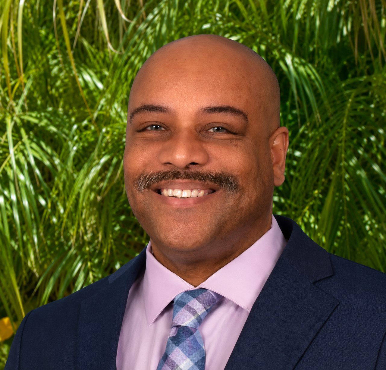 Man in suit smiling in front of green foliage background.